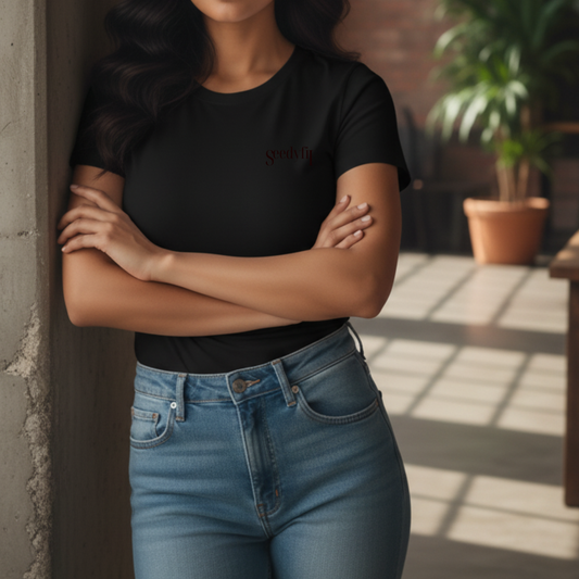Person wearing a black t-shirt and blue jeans standing against a wall with plants in the background.